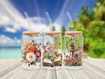 Drei 16 oz Glasbecher mit Bambusdeckel und Glastrinkhalm, dekoriert mit farbenfrohen Motiven aus Blumen, einem aufgeschlagenen Buch und einer Windmühle. Die Becher stehen auf einem Holztisch vor einem Strandhintergrund, was eine entspannte und naturverbun